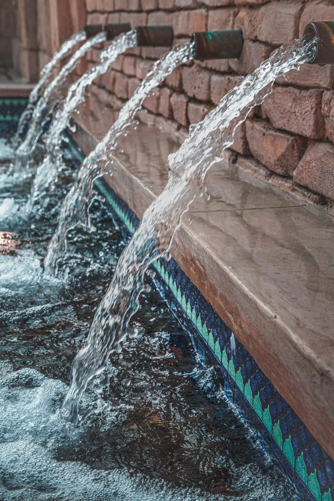 Multiple streams of water flowing from metal spouts into a tiled pool with blue and green mosaic patterns.